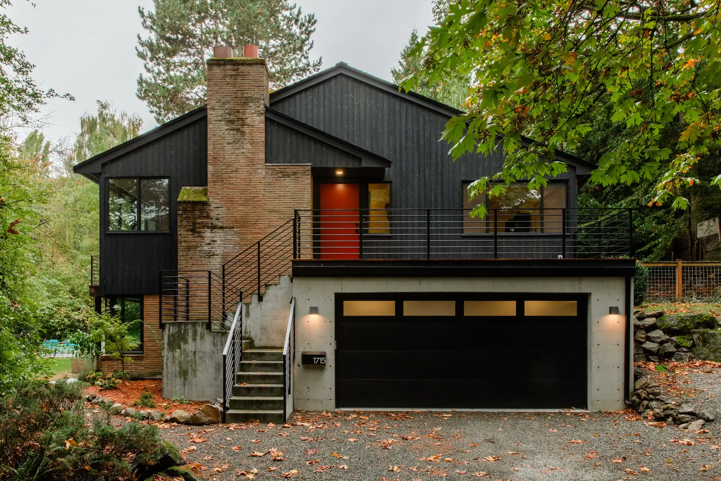Modern black house with brick chimney and garage, surrounded by trees.