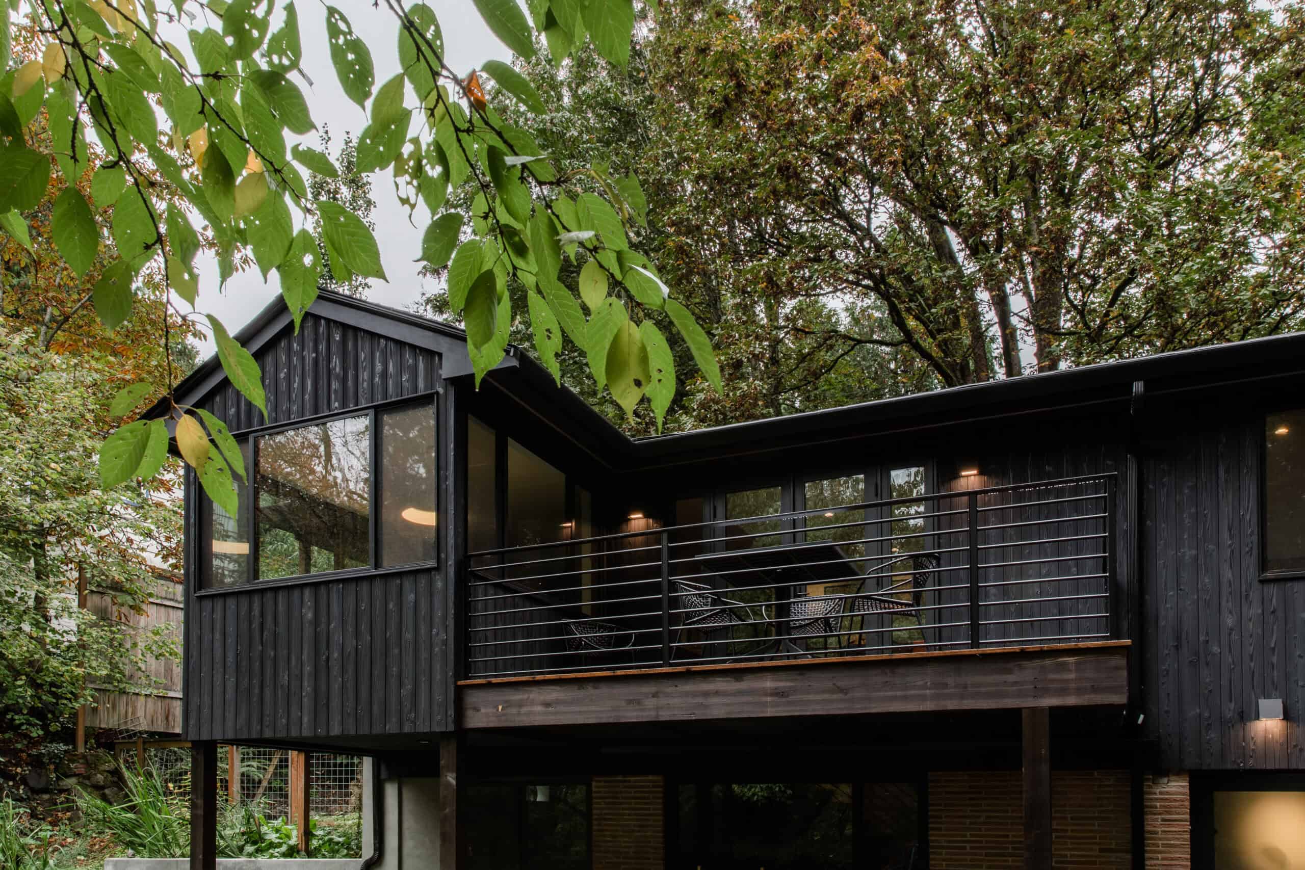 Modern black wooden house with large windows and balcony surrounded by trees.