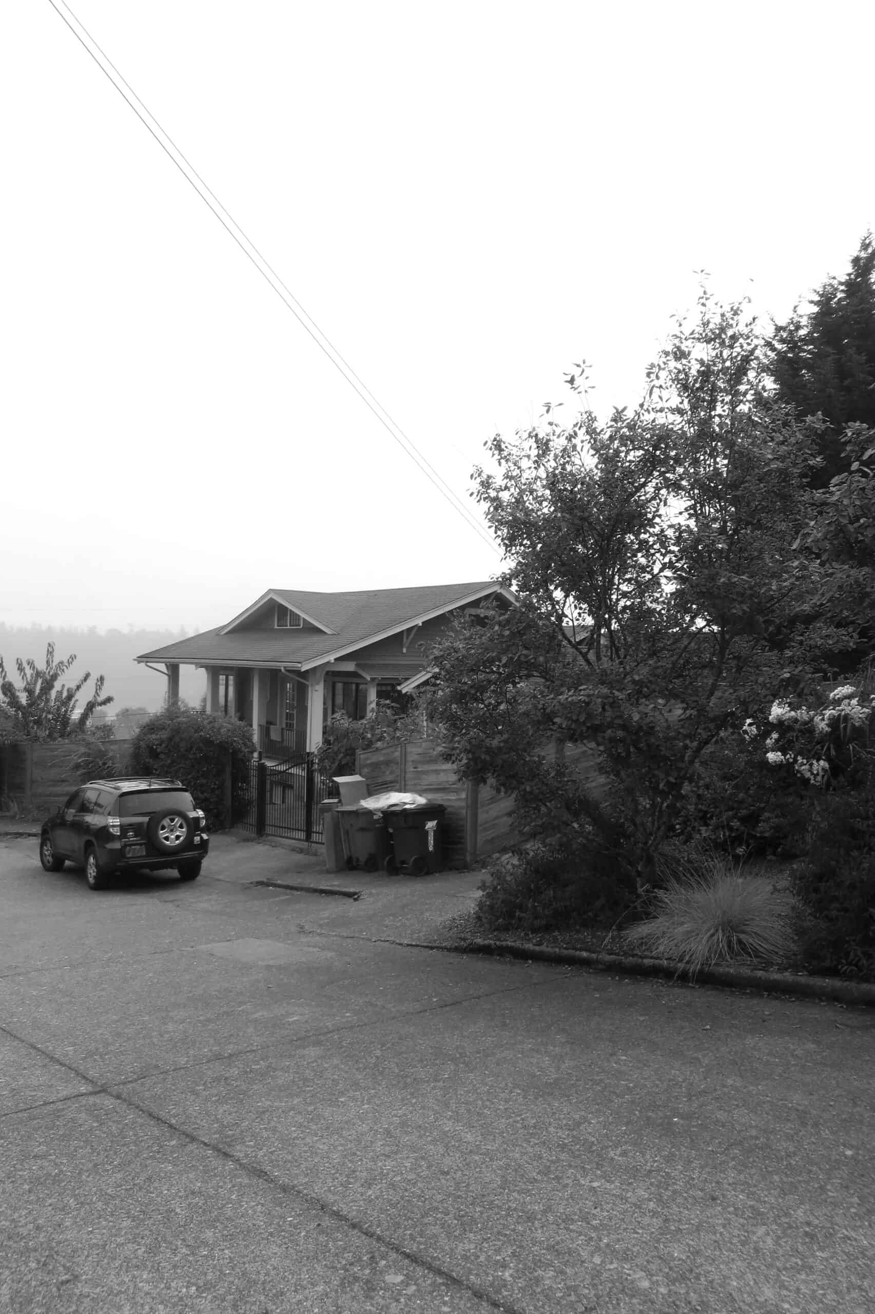 Residential house with a car parked in front and trees nearby in black and white.