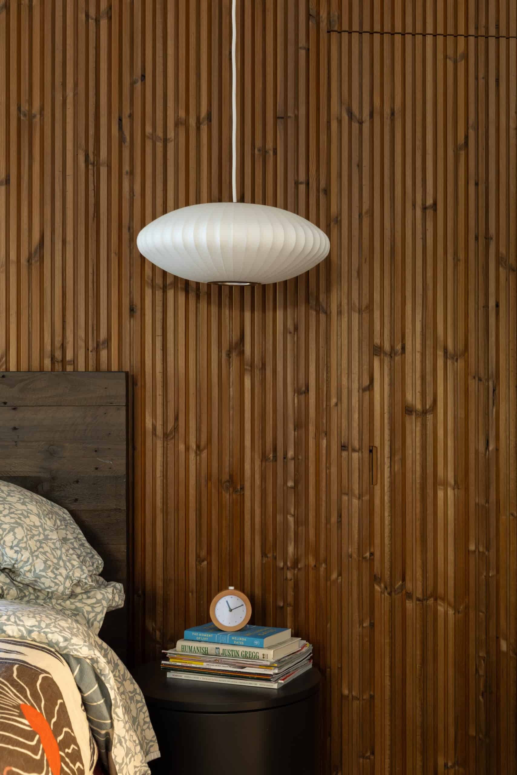 Modern bedroom with wooden wall, pendant light, and bedside table with books and clock.