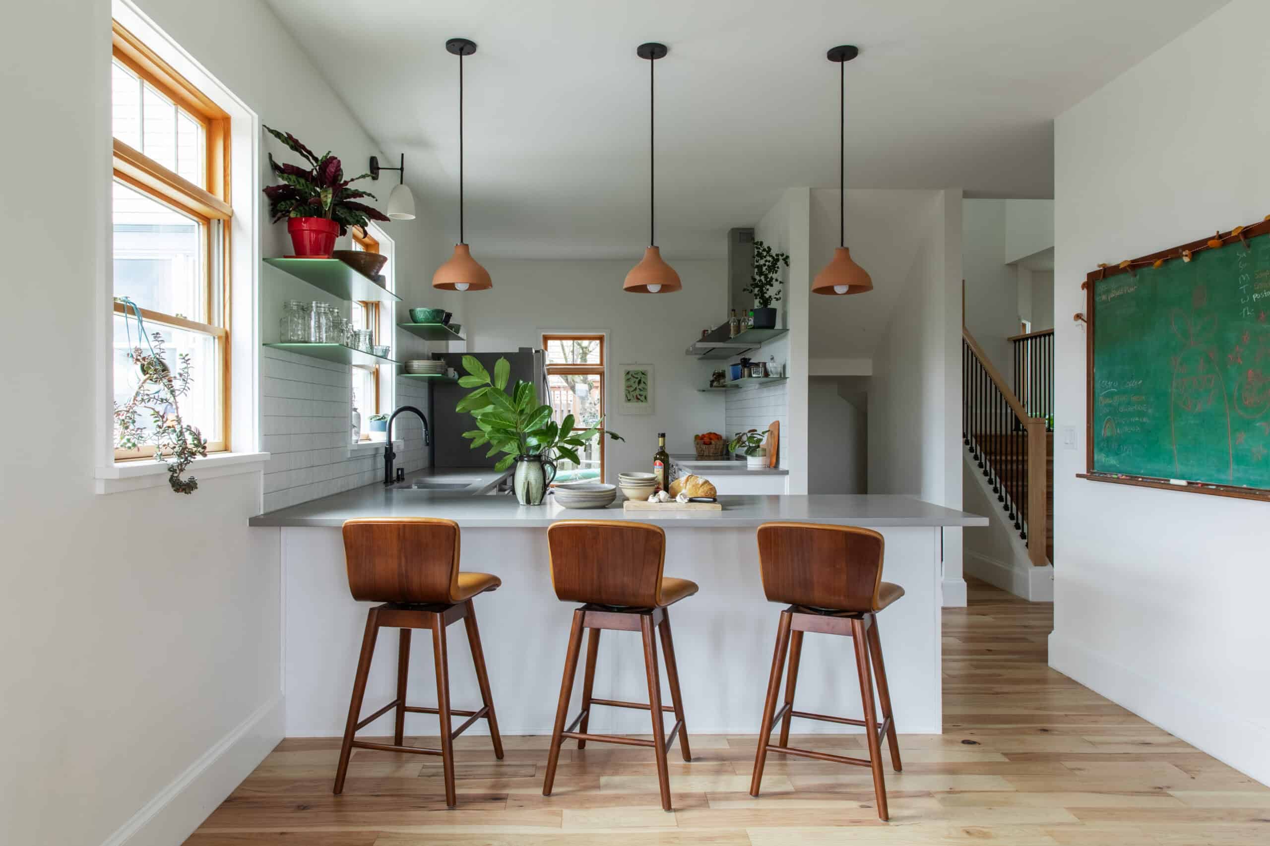 Bright modern kitchen with wooden bar stools, plants, and pendant lighting.