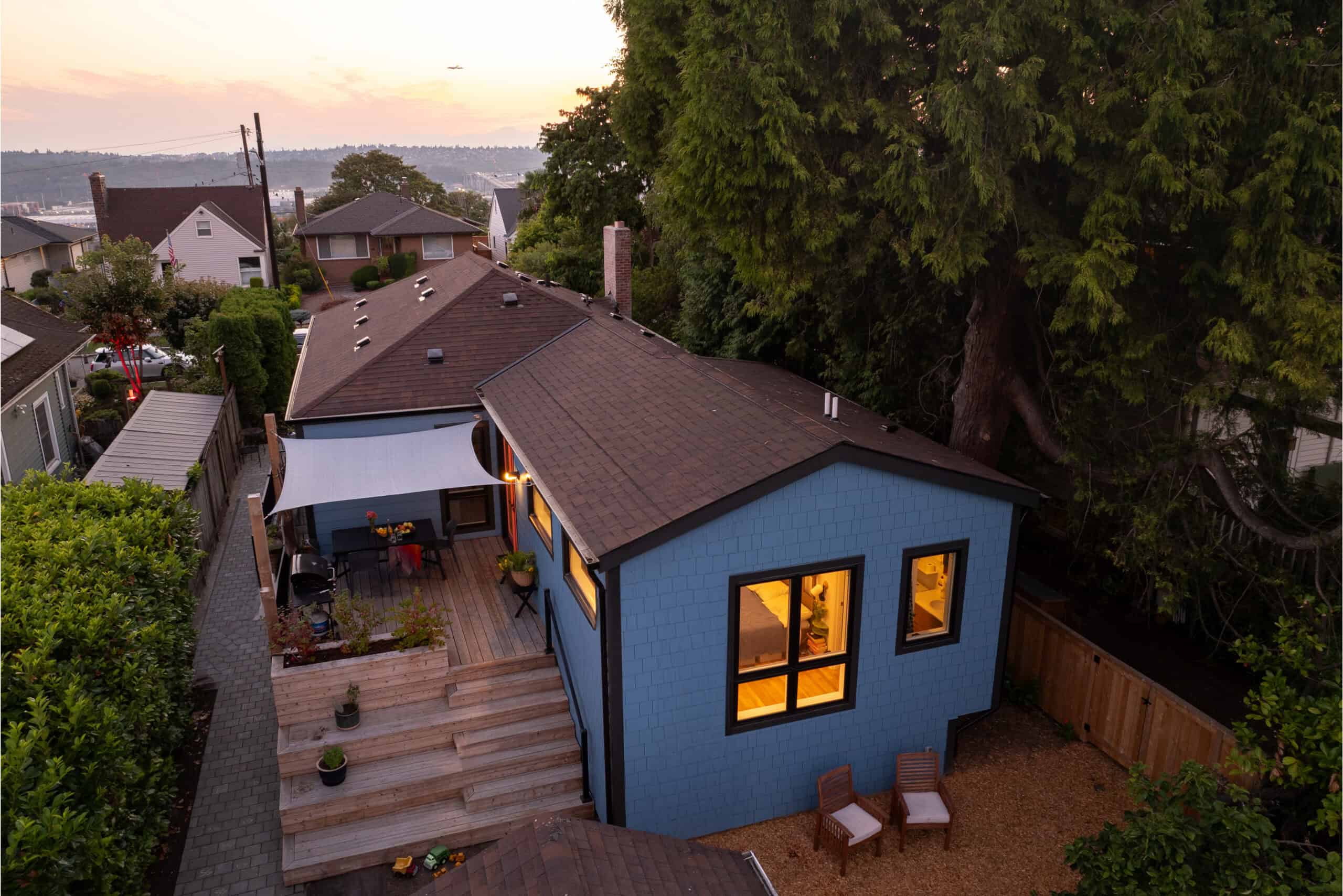 Modern blue house with outdoor deck and large trees in a residential neighborhood.