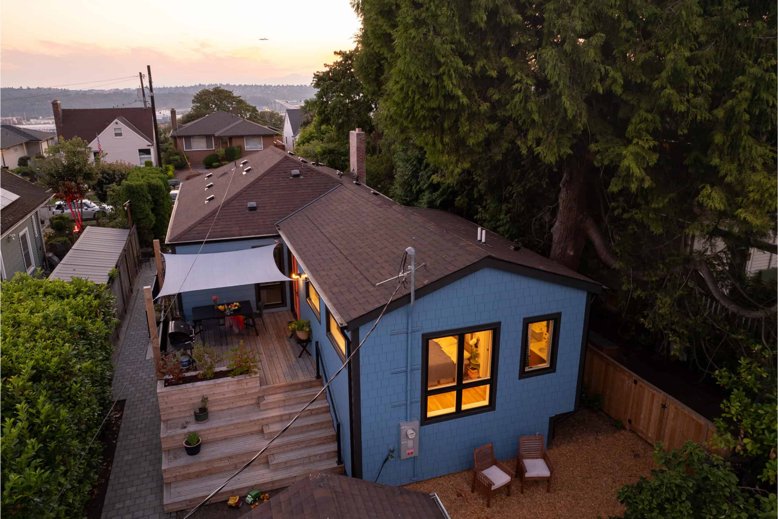 Modern blue house with outdoor deck and large trees in a residential neighborhood.